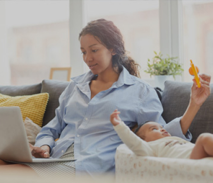 A woman on a couch typing on a laptop with one hand while entertaining her baby with the other hand